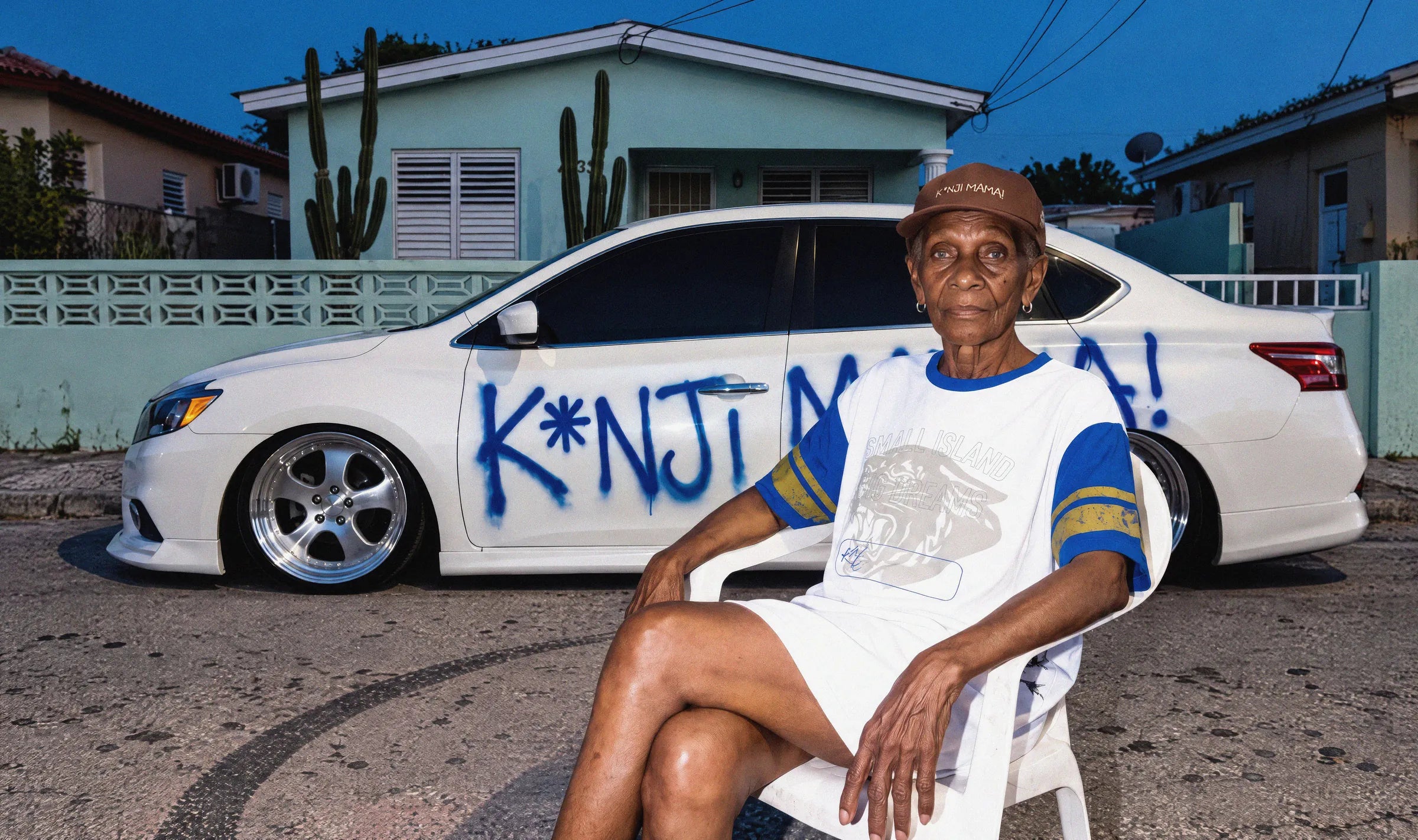 Woman in the 'K*nji Mama' cap and 'Nosta12' T-shirt on a plastic chair in front of a white car sprayed with K*NJI MAMA!